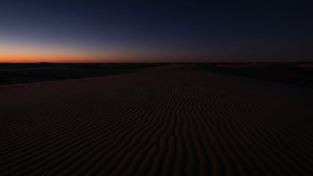 Rippled sand dunes stretch across a desert landscape at twilight, with a dark blue sky and a faint orange glow on the horizon.の素材