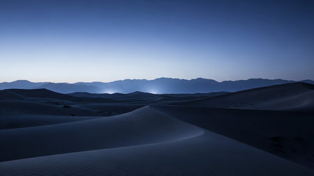 Rolling sand dunes stretch towards distant mountains under a deep blue twilight sky, creating a vast and serene desert landscape.の素材