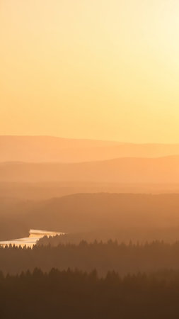 Distant mountain ranges fade into soft hues of orange and yellow at sunset, with a winding river visible through a dark forest.の素材