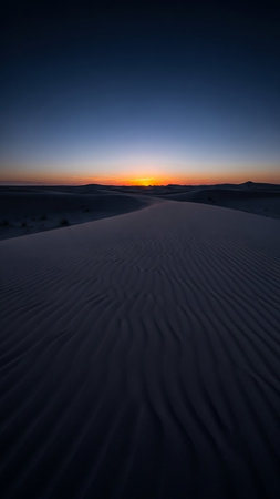 Undulating desert dunes with intricate ripple patterns are bathed in the warm glow of sunset, transitioning to a deep blue twilight sky.の素材