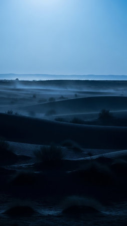 Dark, rolling sand dunes stretch across the landscape under a deep blue twilight sky, with subtle mist creating an ethereal atmosphere.の素材