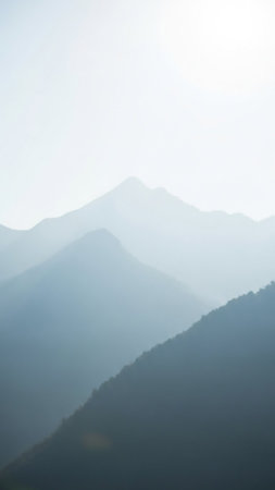 Layers of soft blue mountain peaks fade into a hazy sky, with a dark, forested slope in the foreground under gentle morning light.の素材