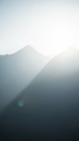 Softly focused mountain silhouettes fade into a bright, hazy sky with a gentle lens flare and subtle green light reflection.の素材