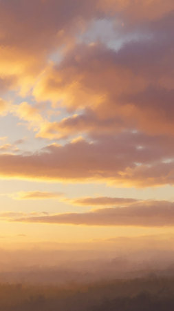 A soft focus view of a sunset sky filled with warm orange, pink, and yellow clouds, with a hazy, indistinct horizon below.の素材