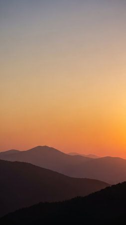 Layered mountain ranges are silhouetted against a vibrant gradient sunset sky transitioning from soft purple to bright orange. A small tower is visible on the highest peak.の素材