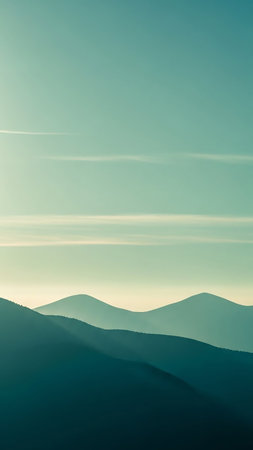 Softly silhouetted teal mountain ridges are layered against a pale, serene sky with subtle horizontal clouds, creating a peaceful landscape.の素材