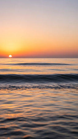 The sun sets over a calm ocean, casting golden and orange light on the gentle waves and the sky. The water reflects the warm hues of the sunset.の素材
