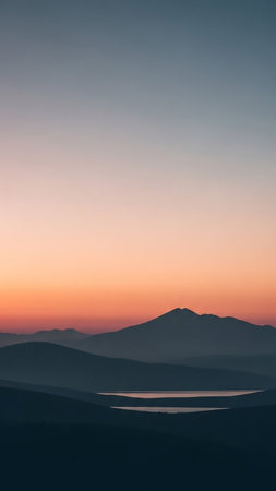Layers of dark mountains and a calm body of water are silhouetted against a gradient sky of orange, pink, and blue at dusk.の素材