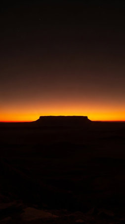 The silhouette of Table Mountain is sharply defined against a vibrant orange and yellow horizon at dusk, with a dark, shadowy foreground.の素材