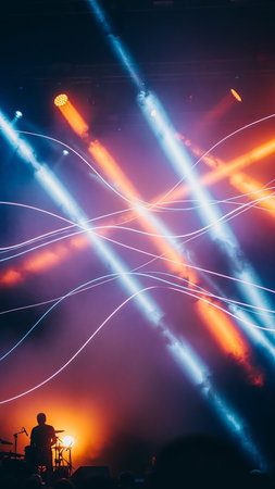 A lone musician's silhouette is visible on a dimly lit stage, bathed in a dramatic display of intersecting blue and orange laser beams and smoke. The vibrant lights create an energetic and dynamic...の素材