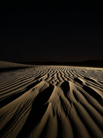 Expansive sand dunes stretch across the frame under a deep black night sky dotted with faint stars. The undulating surface of the sand is highlighted by dramatic shadows, creating a stark and...の素材