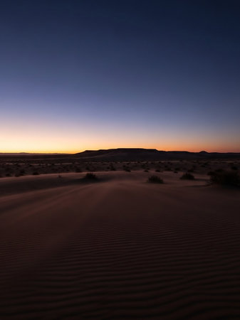 A vast desert landscape at twilight undulating sand dunes with wind-swept ripples in the foreground, leading to a silhouetted mountain range on the horizon under a gradient of deep blue and orange...の素材