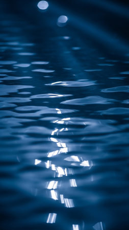 The dark blue surface of water is illuminated by bright, abstract light reflections. Ripples and waves create a dynamic, shimmering pattern with a moody, serene atmosphere.の素材