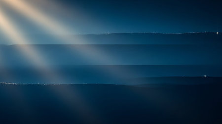 Soft, ethereal light rays cut through layered dark blue mist, illuminating faint distant city lights, creating a moody and atmospheric scene.の素材