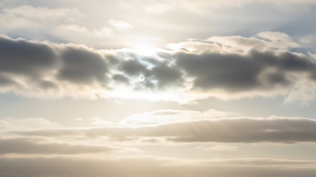The bright sun shines through layered white and gray clouds, casting visible sunbeams and a soft, diffused light across the expansive sky.の素材