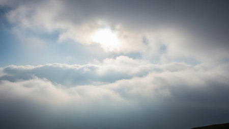 The bright sun shines through a layer of thick, soft clouds, casting a diffused light against a backdrop of blue sky.の素材