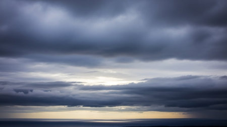 Dark, stormy grey clouds dominate the sky above a calm sea, with a faint yellow horizon and subtle light breaking through the dense cloud cover.の素材