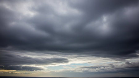 Dark, heavy storm clouds dominate the sky above a calm sea horizon, with thin strips of pale blue sky visible through the dense overcast.の素材
