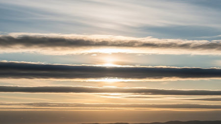 Horizontal layers of dark gray and light blue clouds are intersected by the sun peeking through the middle, creating a striking atmospheric horizon.の素材