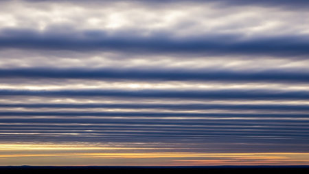 Across showing horizontal bands of dark gray and blue clouds stretch across the sky above a thin strip of orange and yellow sunset light and a dark horizon line...の素材