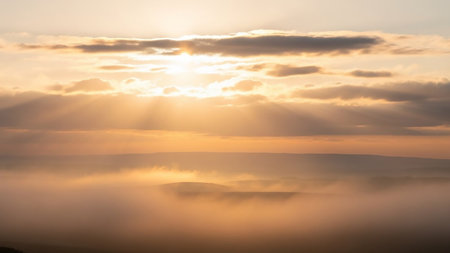 Golden sunrise with sunbeams piercing through clouds over a misty landscape of rolling hills, bathed in warm orange light.の素材