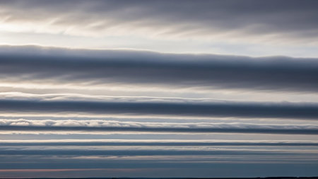 Horizontal layers of wispy gray and white clouds are illuminated by soft sunlight, creating a textured sky backdrop with a subtle horizon line.の素材