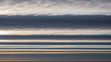 Horizontal layers of gray and white clouds with thin bright edges create a striped atmospheric pattern against a pale sky.の素材
