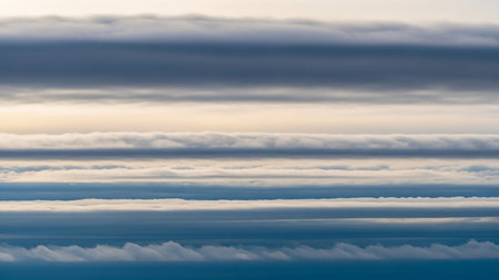 Horizontal layers of stratocumulus clouds in varying shades of gray blue and white create a textured pattern against a soft golden light.の素材