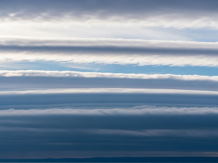 Above showing layered stratus clouds in shades of blue and white creating horizontal bands across a bright sky above a dark horizon line with subtle hints of...の素材