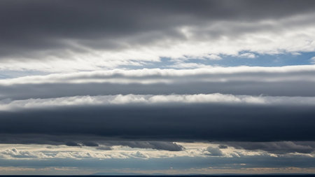 Horizontal bands of dark grey and white clouds are set against a bright blue sky, with distant land visible below.の素材