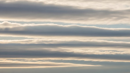 Horizontal layers of clouds are illuminated by soft sunlight, creating a gentle texture and subtle play of light and shadow across the sky.の素材
