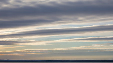 Horizontal layers of grey, white, and pale blue clouds create a serene and vast sky above a dark, calm horizon line, suggesting a peaceful natural scene.の素材