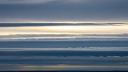 Subtle showing horizontal layers of dark blue and grey clouds are separated by thin bands of soft yellow and white light, creating a textured sky panorama....の素材
