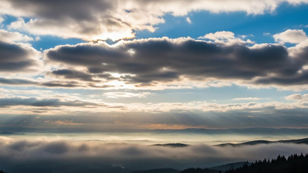 A mountain landscape shrouded in fog with sunbeams piercing through scattered clouds and a clear blue sky above.の素材