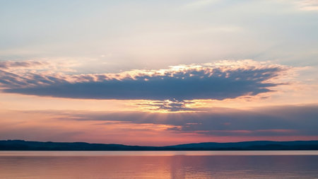 Silhouetted showing serene sunset over a calm lake with silhouetted hills and sun rays breaking through dramatic clouds reflected in the water. resolution...の素材