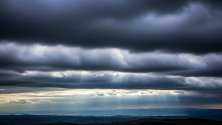Dark showing rays of sunlight piercing through dark, heavy storm clouds over a distant landscape, creating a dramatic and hopeful contrast of light and shadow....の素材