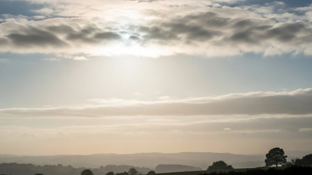 Natural showing rolling hills and trees under a bright, hazy sky with the sun partially obscured by soft, layered clouds creating a gentle landscape scene keywords:...の素材