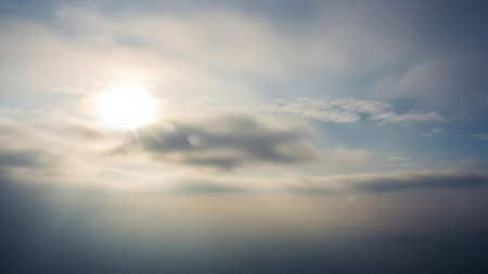 Bright sun illuminates a hazy sky with scattered clouds and a soft lens flare effect creating a diffused atmospheric perspective.の素材