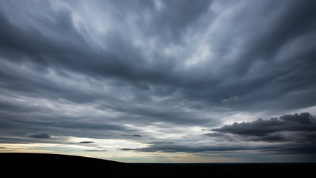 Dark, heavy storm clouds dominate the sky, casting a dramatic mood over a silhouetted landscape with a faint line of light on the horizon.の素材
