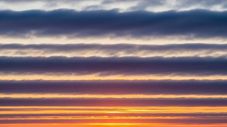 Horizontal layers of clouds display vibrant sunset colors including orange, purple, and blue, with a bright sun glow.の素材