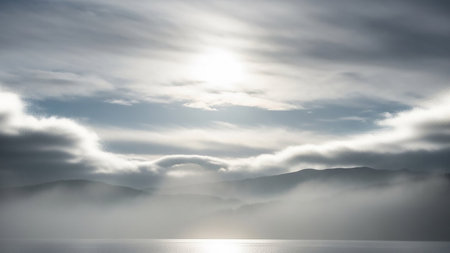 A bright sun shines through a cloudy sky above misty mountain ranges and a calm, reflective body of water.の素材