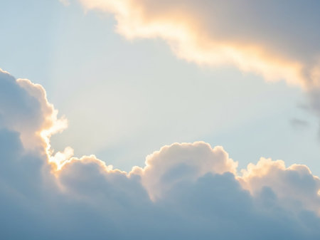 Soft, diffused sunlight casts a warm golden glow on the edges of puffy white clouds against a serene pale blue sky.の素材