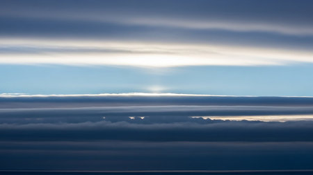 Dramatic layered clouds in shades of dark blue and gray are illuminated by a bright white light breaking through the atmospheric horizon above the sea.の素材