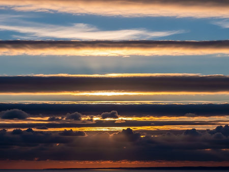 Dramatic horizontal cloud layers in shades of orange, gold, gray, and dark blue are intersected by sunbeams, creating a striking sunset sky backdrop.の素材
