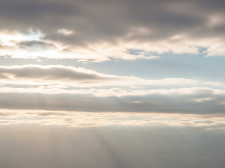 Sun rays pierce through layered clouds, casting dramatic light beams across a muted sky during sunrise or sunset.の素材