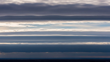 Horizontal layers of grey clouds with bright white edges create a stratified sky pattern. Sunlight illuminates the cloud formations above a dark horizon.の素材
