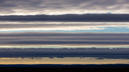 Horizontal layers of dark gray and white clouds create a striped pattern across a pale blue sky above a soft orange and dark horizon.の素材
