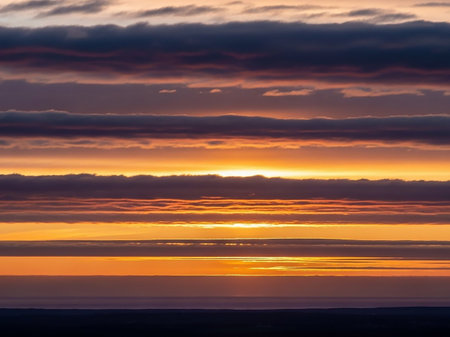 Orange showing horizontal layers of dark grey and vibrant orange clouds creating a dramatic striped pattern across the sky at sunset over a calm body of water.の素材