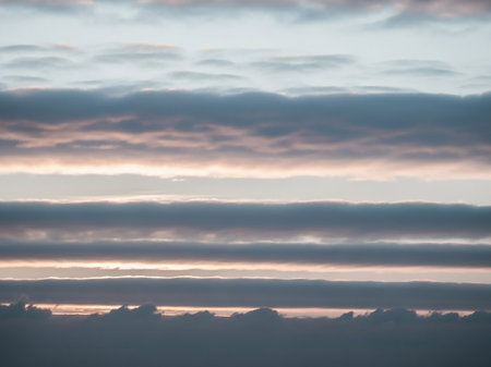 Horizontal layers of soft grey and pale pinkish clouds fill the sky at dawn, with subtle sunlight illuminating the gaps between the formations.の素材