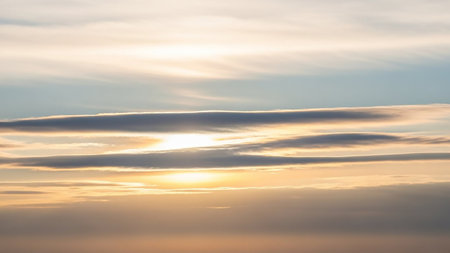 Rays showing soft golden sunlight breaks through horizontal layers of wispy clouds and a pale blue sky creating a serene and tranquil atmospheric horizon. keywords:...の素材